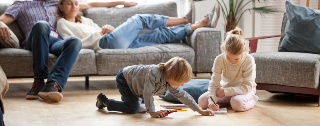 two adults sitting on couch with two children playing on floor in living room