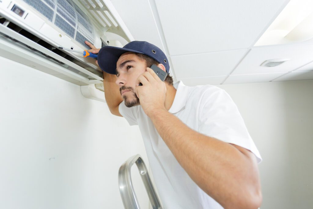 young man wearing blue hat on ladder holding phone to ear calling about hvac noise repair