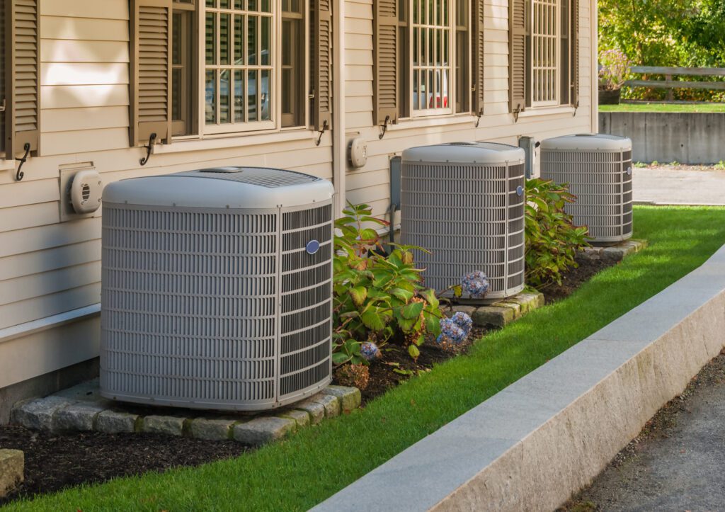 Three industrial HVAC units against wall with grass and plants around